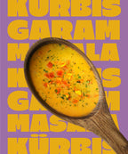 Ein Holzlöffel in dem Miss Mineva's fermentierte Suppe mit Geschmacksrichtung Kürbis-Garam-Masala vor einem bunten Hintergrund zu sehen ist